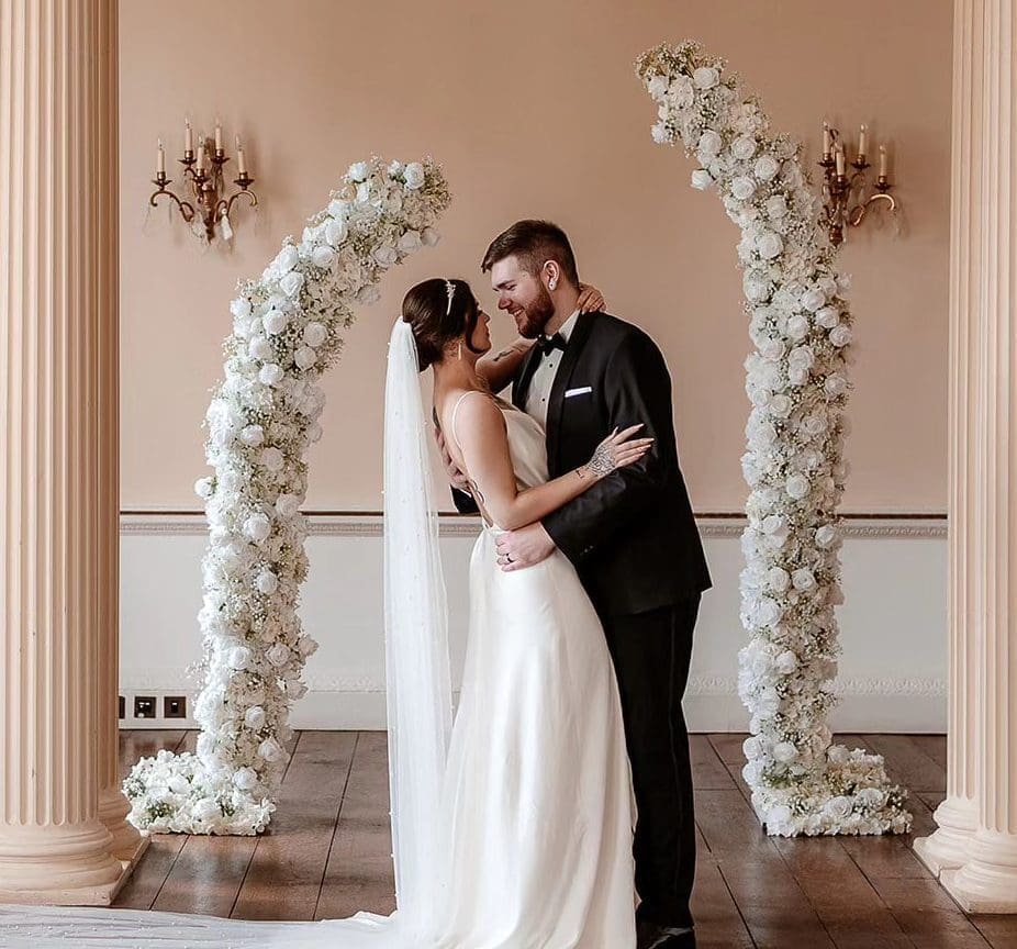 Wedding couple in embrace by wedding arch of silk flowers from The Floral hire